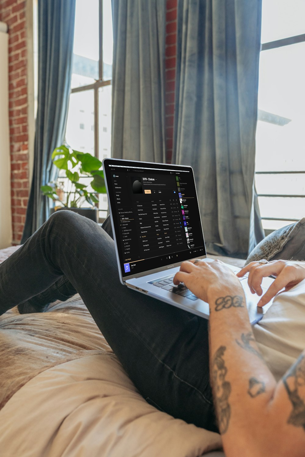 person in gray sweater and blue denim jeans sitting on brown bed using black laptop computer