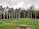 A well-maintained garden with a neatly trimmed green lawn is bordered by a row of tall palm trees. The garden area is surrounded by various shades of green foliage and flowering shrubs, creating a serene and natural setting. There is a wooden bench in the foreground, and the sky appears overcast with a hint of grey clouds.