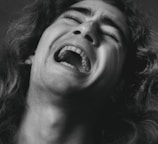A close-up black and white photograph of a person experiencing a strong emotion, likely during an intense moment or expression of agony. The person's mouth is open wide, eyes are partially closed, and the features are dramatically lit, highlighting the texture of the skin and hair.
