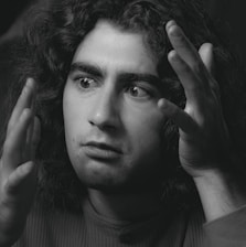A black and white portrait of a person with long curly hair and expressive eyes. The person appears to be deep in thought or conversation, with hands raised near the face. The lighting highlights the facial features, casting subtle shadows.