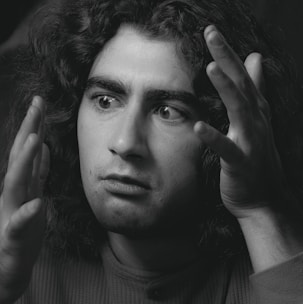 A black and white portrait of a person with long curly hair and expressive eyes. The person appears to be deep in thought or conversation, with hands raised near the face. The lighting highlights the facial features, casting subtle shadows.
