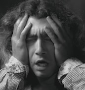 A person with long hair is holding their head with both hands, eyes closed, and mouth slightly open, displaying an expression of distress or frustration.