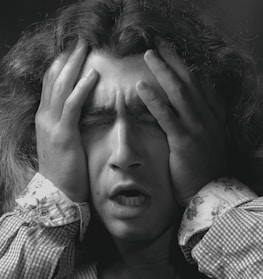A person with long hair is holding their head with both hands, eyes closed, and mouth slightly open, displaying an expression of distress or frustration.