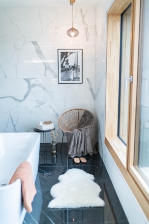 A modern bathroom with marble walls and a glossy black floor. There is a free-standing white bathtub with a pink towel draped over the edge. A cozy corner features a wicker chair covered with a gray blanket and a pair of slippers on the floor. A small side table holds a stack of books and decorative items in a vase. A black and white framed photo is mounted on the wall, and a single light fixture hangs above. A large window allows natural light to brighten the space.