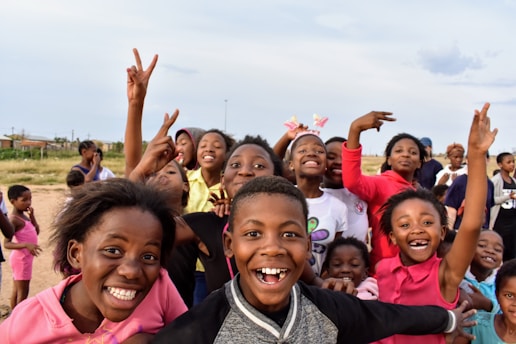 A group of smiling and excited children outdoors, many of whom are making playful gestures and showing expressions of joy. The background shows a field and a few scattered houses, suggesting a rural setting.