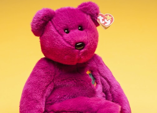 A bright pink plush teddy bear sits against a yellow background. The bear has a soft, fuzzy texture with black eyes and a black nose. A heart-shaped tag with a logo hangs from its ear, and an embroidered design on its chest adds detail.