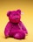 A bright pink plush teddy bear sits against a yellow background. The bear has a soft, fuzzy texture with black eyes and a black nose. A heart-shaped tag with a logo hangs from its ear, and an embroidered design on its chest adds detail.