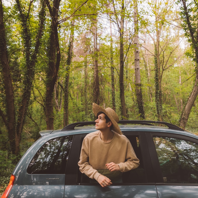 A guy wearing a hat looking through a window watching a beautiful green natural world around him
