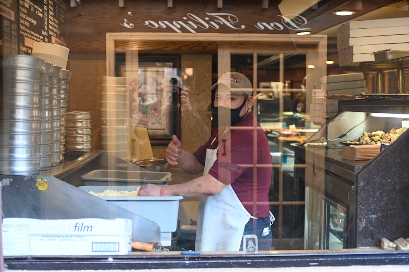 A person wearing a mask and apron stands inside a pizzeria, surrounded by pizza boxes and trays of food. They are giving a thumbs-up gesture through a window, with stacks of pizza pans and a counter with various ingredients visible. The interior includes warm lighting and wooden fixtures.