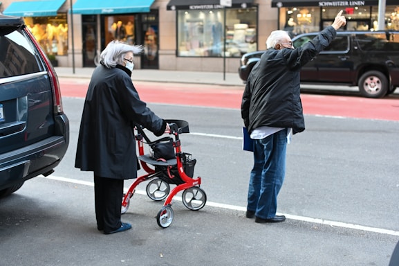 An elderly woman with gray hair, wearing a mask and a black coat, stands on the sidewalk with a red walker. Next to her, an older man dressed in a black jacket and jeans is raising his hand to hail a cab. The scene takes place on a city street with a row of shops in the background.