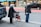 An elderly woman with gray hair, wearing a mask and a black coat, stands on the sidewalk with a red walker. Next to her, an older man dressed in a black jacket and jeans is raising his hand to hail a cab. The scene takes place on a city street with a row of shops in the background.