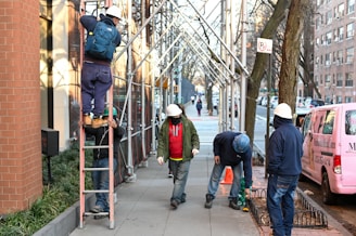 A group of construction workers wearing safety helmets are working on a city sidewalk next to a building. One worker is climbing a ladder with a backpack, another is standing on the ground holding the ladder, and two workers are engaged in a task on the pavement. A pink van is parked on the street beside them. Scaffolding lines the sidewalk and trees are visible along the street.