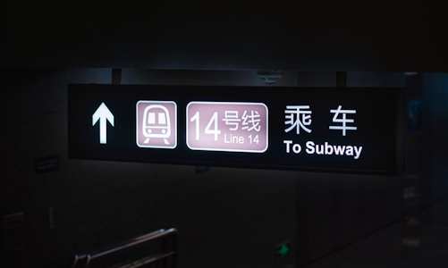 An overhead illuminated sign in a subway station displays directional information. It features an arrow pointing upward, an icon of a train, the text 'Line 14' with both Chinese and English translations, and the words 'To Subway' in clear white lettering on a black background.