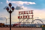 A large neon sign reading 'PUBLIC MARKET' stands prominently against a bright sky with scattered clouds. A street lamp with round glass covers and a Ferris wheel are visible in the background. The scene is urban, capturing the essence of a bustling marketplace.