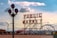 A large neon sign reading 'PUBLIC MARKET' stands prominently against a bright sky with scattered clouds. A street lamp with round glass covers and a Ferris wheel are visible in the background. The scene is urban, capturing the essence of a bustling marketplace.