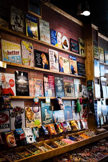 The image depicts a colorful store display featuring an array of vintage-style metal signs and candy packets. The signs display various retro illustrations, humorous quotes, and nostalgic icons. Below the signs, the shelves hold an assortment of bagged candies and snacks, organized in wooden compartments.
