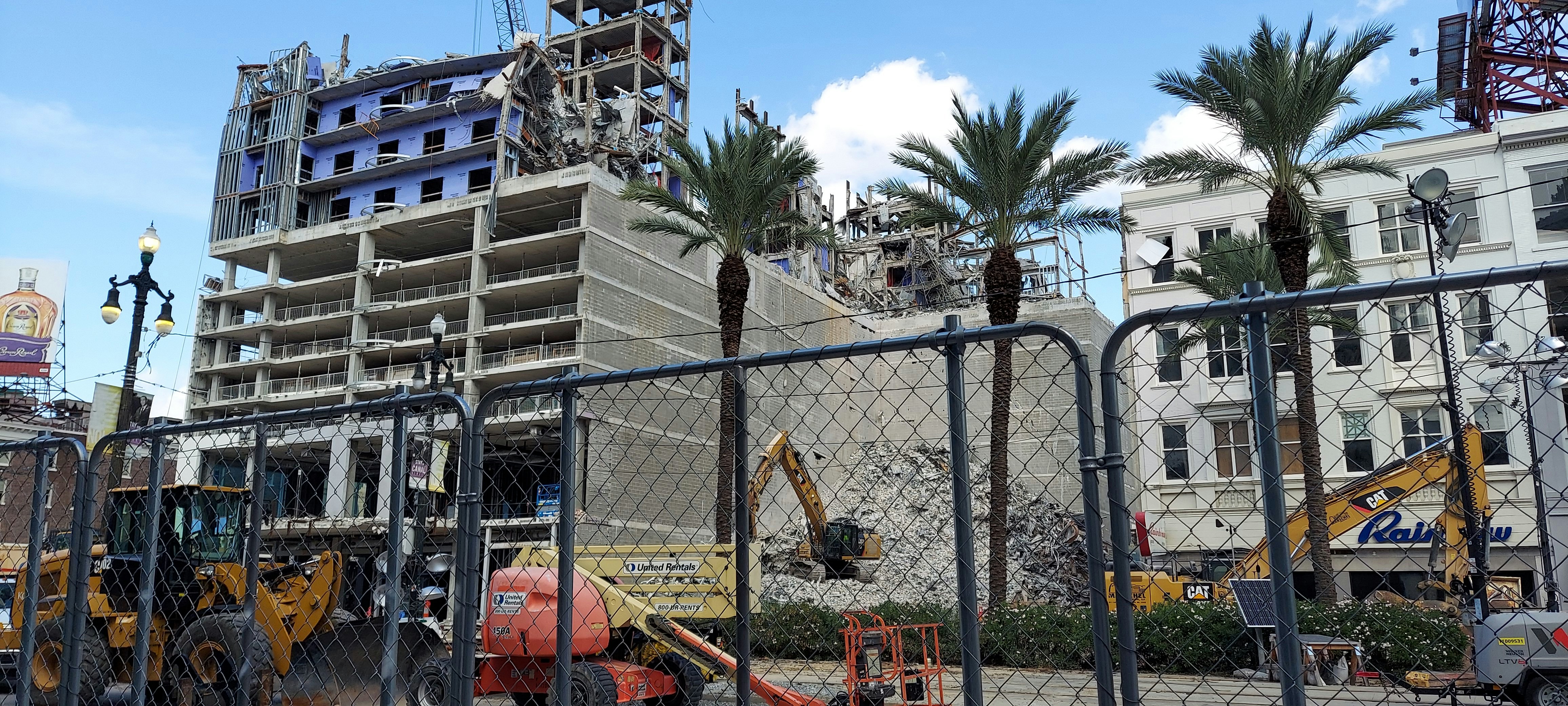 A partially demolished building with exposed floors and structural elements is visible behind a chain-link fence. Construction machinery, including excavators and a lift, are actively working on the site. Palm trees are standing in front of the building, and a billboard is visible in the background under a partly cloudy sky.