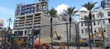 A partially demolished building with exposed floors and structural elements is visible behind a chain-link fence. Construction machinery, including excavators and a lift, are actively working on the site. Palm trees are standing in front of the building, and a billboard is visible in the background under a partly cloudy sky.