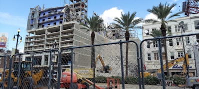 A partially demolished building with exposed floors and structural elements is visible behind a chain-link fence. Construction machinery, including excavators and a lift, are actively working on the site. Palm trees are standing in front of the building, and a billboard is visible in the background under a partly cloudy sky.