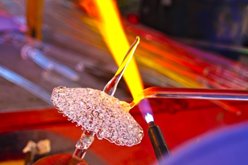 A detailed view of glassblowing with a focused flame shaping molten glass, creating intricate textures and patterns. Bright, vivid colors contrast the delicate transparency of the glass.