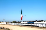 A large flagpole displays a Mexican flag prominently against a clear blue sky. Below the flag, there's a wide canal or channel lined with concrete, bordered by fences. In the background, there are commercial buildings and a parking lot. Trees and streetlights are scattered along the horizon.