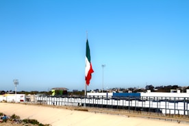 A large flagpole displays a Mexican flag prominently against a clear blue sky. Below the flag, there's a wide canal or channel lined with concrete, bordered by fences. In the background, there are commercial buildings and a parking lot. Trees and streetlights are scattered along the horizon.