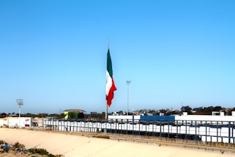 A large flagpole displays a Mexican flag prominently against a clear blue sky. Below the flag, there's a wide canal or channel lined with concrete, bordered by fences. In the background, there are commercial buildings and a parking lot. Trees and streetlights are scattered along the horizon.