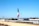 A large flagpole displays a Mexican flag prominently against a clear blue sky. Below the flag, there's a wide canal or channel lined with concrete, bordered by fences. In the background, there are commercial buildings and a parking lot. Trees and streetlights are scattered along the horizon.