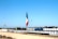 A large flagpole displays a Mexican flag prominently against a clear blue sky. Below the flag, there's a wide canal or channel lined with concrete, bordered by fences. In the background, there are commercial buildings and a parking lot. Trees and streetlights are scattered along the horizon.