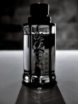 A sleek, reflective glass bottle stands upright against a blurred, dark background. The bottle is cylindrical with embossed text on the surface, and it features a prominent black cap. The lighting creates a dramatic and elegant effect, highlighting the contours and texture of the bottle.