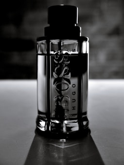 A sleek, reflective glass bottle stands upright against a blurred, dark background. The bottle is cylindrical with embossed text on the surface, and it features a prominent black cap. The lighting creates a dramatic and elegant effect, highlighting the contours and texture of the bottle.