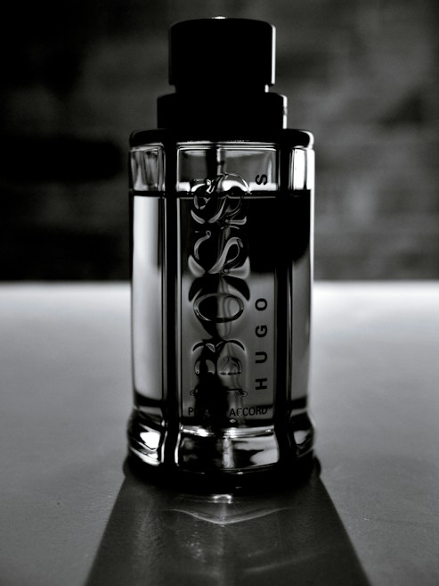 A sleek, reflective glass bottle stands upright against a blurred, dark background. The bottle is cylindrical with embossed text on the surface, and it features a prominent black cap. The lighting creates a dramatic and elegant effect, highlighting the contours and texture of the bottle.