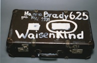 A vintage brown suitcase with visible wear has white painted lettering on its surface. The text includes a name, date of birth, and the word 'Waisenkind', which translates to 'orphan' in German. The suitcase features metal locks and protective corners.