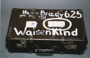 A vintage brown suitcase with visible wear has white painted lettering on its surface. The text includes a name, date of birth, and the word 'Waisenkind', which translates to 'orphan' in German. The suitcase features metal locks and protective corners.