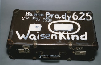 A vintage brown suitcase with visible wear has white painted lettering on its surface. The text includes a name, date of birth, and the word 'Waisenkind', which translates to 'orphan' in German. The suitcase features metal locks and protective corners.