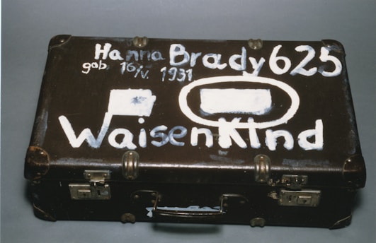 A vintage brown suitcase with visible wear has white painted lettering on its surface. The text includes a name, date of birth, and the word 'Waisenkind', which translates to 'orphan' in German. The suitcase features metal locks and protective corners.