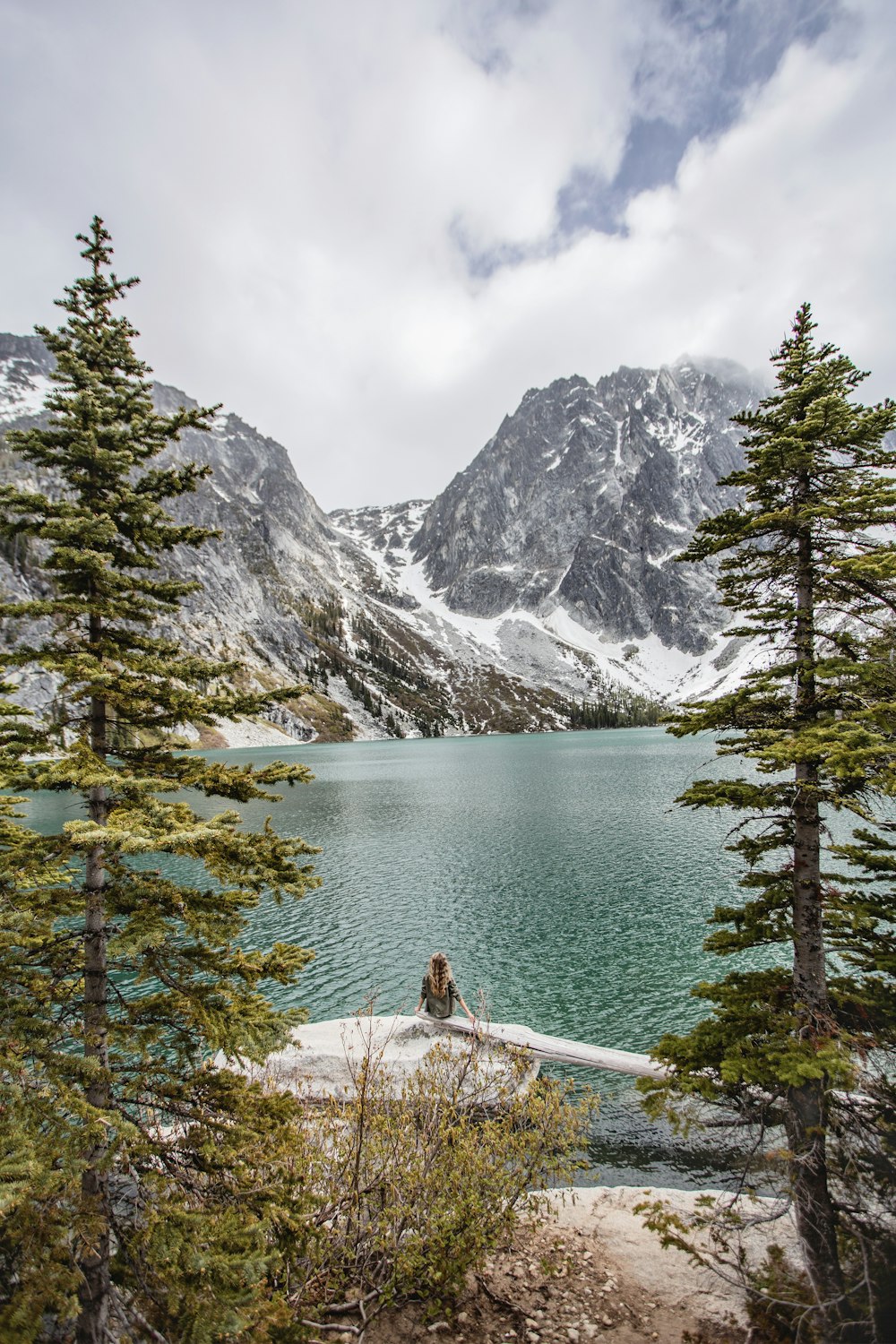 Persona che si siede sul molo di legno marrone vicino al lago e alla montagna coperta di neve durante il giorno