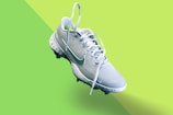 A light blue athletic shoe with a prominent white swoosh symbol and white laces is centered on a vibrant, two-toned green background. The shoe features textured detailing on the sole and upper fabric, indicating it's designed for sports.