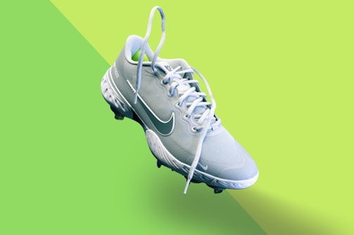 A light blue athletic shoe with a prominent white swoosh symbol and white laces is centered on a vibrant, two-toned green background. The shoe features textured detailing on the sole and upper fabric, indicating it's designed for sports.