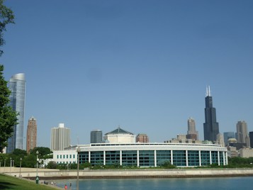 A skyline with modern skyscrapers including a distinctive black tower with antennae. A rounded, low white building with a green, manicured lawn stretches along a waterfront in the foreground. A few people walk along the promenade beside the water, and lush green trees partially frame the scene on the left side.