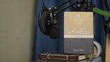 A book with Arabic text on its cover is propped up against a microphone setup. The book is placed on top of another book, and there is a green plant leaf partially visible. The background features a blue curtain and a beige wall.