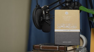 A book with Arabic text on its cover is propped up against a microphone setup. The book is placed on top of another book, and there is a green plant leaf partially visible. The background features a blue curtain and a beige wall.
