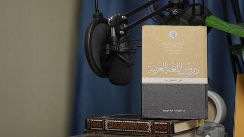 A book with Arabic text on its cover is propped up against a microphone setup. The book is placed on top of another book, and there is a green plant leaf partially visible. The background features a blue curtain and a beige wall.