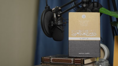 A book with Arabic text on its cover is propped up against a microphone setup. The book is placed on top of another book, and there is a green plant leaf partially visible. The background features a blue curtain and a beige wall.