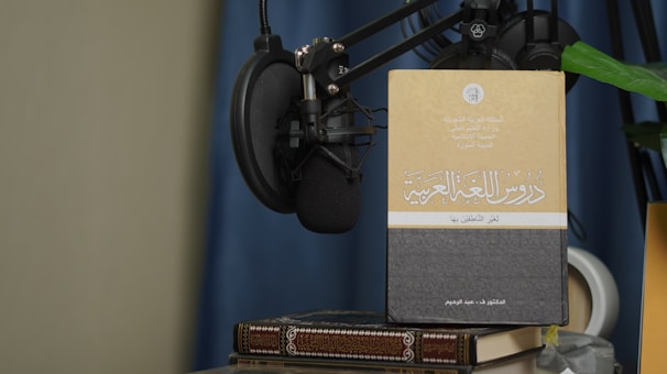 A book with Arabic text on its cover is propped up against a microphone setup. The book is placed on top of another book, and there is a green plant leaf partially visible. The background features a blue curtain and a beige wall.