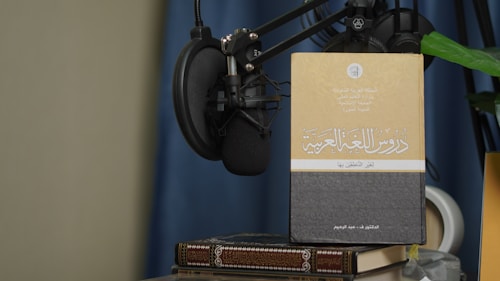 A book with Arabic text on its cover is propped up against a microphone setup. The book is placed on top of another book, and there is a green plant leaf partially visible. The background features a blue curtain and a beige wall.