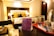 A hotel room with a neatly made bed featuring two yellow cushions and two bedside lamps providing warm lighting. A black and white headboard contrasts with the beige walls. In the foreground, a tray on a counter holds glass tumblers, mugs, and various coffee sachets. A promotional card for travel assistance leans against the tray.