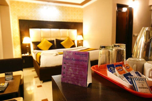 A hotel room with a neatly made bed featuring two yellow cushions and two bedside lamps providing warm lighting. A black and white headboard contrasts with the beige walls. In the foreground, a tray on a counter holds glass tumblers, mugs, and various coffee sachets. A promotional card for travel assistance leans against the tray.
