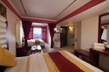 A hotel room with two beds, adorned with red and yellow bedding and pillows. The room features red curtains, a large window with a view of a mountainous landscape, and a wall-mounted television above a small cabinet. There is a vanity with a mirror, folded towels, and toiletries on the right side. Recessed lighting and an illuminated wall sconce provide lighting, and the overall color scheme is red and white.