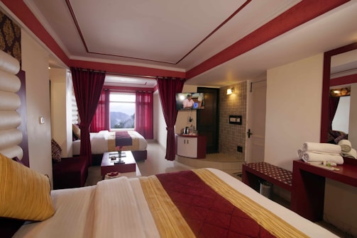 A hotel room with two beds, adorned with red and yellow bedding and pillows. The room features red curtains, a large window with a view of a mountainous landscape, and a wall-mounted television above a small cabinet. There is a vanity with a mirror, folded towels, and toiletries on the right side. Recessed lighting and an illuminated wall sconce provide lighting, and the overall color scheme is red and white.